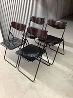 Front and side view of four black IKEA folding chairs arranged in a group on a grey floor in a warehouse setting.