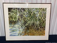 Framed watercolor print showing water with reflections and green plants, overall view including frame.