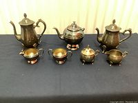 Full set of seven silverplated tea service pieces displayed on dark cloth, showing tarnishing and the overall condition including broken handles.