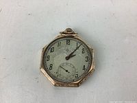 Front view of vintage octagonal pocket watch showing dial with Arabic numerals, short minute and hour hands, subsidiary seconds dial, gold filled engraved bezel, and missing glass crystal.