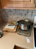 Photo of countertop showing two wooden cutting boards stacked next to two oven mitts and a large stainless steel cooking pot with lid, placed on top of three glass Pyrex baking dishes.