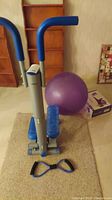 Full view of the Wagan Tech stepper with blue handlebars and pedals, purple Gaiam balance ball in background, and resistance band attachments on the floor in front.