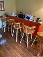 Five solid wood bar stools positioned against a red bar counter on a hardwood floor, showing full view of round seats, backrests, and metal footrests.