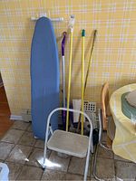 Photo showing a blue ironing board, white step stool, four mops with different colored handles, and a yellow watering can against a yellow floral wallpaper background.