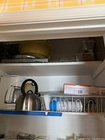 Two shelves showing various kitchen items including a white drip coffee maker, stainless steel kettle, white dish drainer with cups, aluminum baking trays, and a yellow mixing bowl on the top shelf