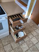 Three kitchen drawers open showing organized kitchen utensils and cutlery including spoons, forks, knives, and various kitchen gadgets.