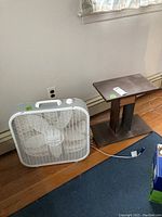 Photo showing wooden TV stand and white Lasko box fan side by side on hardwood floor near wall and baseboard heating.