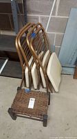 Four metal folding chairs stacked with beige padded seats and backs, showing rust and wear, next to a worn wicker stool.