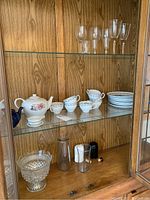 Three shelves in wooden China cabinet showing glassware and china including teapots, cups, plates, vases, salt and pepper shakers