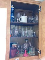 Open kitchen cabinet showing three shelves with various glassware, mugs, drinking glasses, shot glasses, and a blue Pyrex water bottle on top shelf.