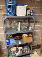 Metal shelving with various vintage glassware including vases, pitchers, wine glasses, ceramic salt and pepper shakers, small jars, bowls, a white cooler, and some cleaning spray cans on top shelf.