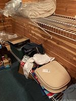 View of shelf with folded sweaters, white gloves, boxes, linens and stacked beige seat pads in plastic wrapping.