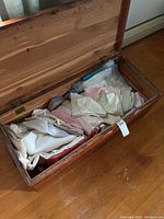 Open wooden chest containing assorted folded linens, blankets, and other items on a wood floor.