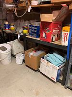 Metal shelving unit with plywood shelves in basement area, holding several packed boxes and miscellaneous items, showing condition and shelving materials.