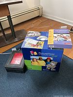 Shows Epson PictureMate Snap box and black photo storage box on carpet near window baseboard.