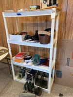 White plastic shelving unit with four shelves seen against wood-paneled wall, unit shows some light dust and dirt, measurements approx 34x15x56 inches, contains box and miscellaneous items not included in sale.