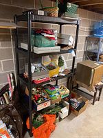 Full view of black plastic four-tier storage shelf unit with various stored items and boxes, against a concrete block basement wall.
