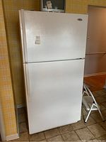 Front and side view of the white Whirlpool top freezer refrigerator placed in kitchen with yellow plaid wallpaper, showing door handles and overall condition.