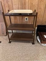 Metal shelf unit with three wooden shelves and collapsible plastic tray on top, against wood panel wall.