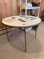 Round white vinyl top card table with metal folding legs, shown from an angle with some paperwork on top on beige carpet.