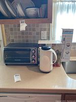 Black & Decker toaster oven, white thermos kettle, Invitations steamer set box, and Cuisinart hand blender box arranged on kitchen counter with dishes above