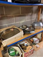 Basement shelf showing various silver plate trays and bowls along with the metal roaster lid partly visible, items arranged for sale in storage area.