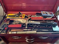 Full top view of open red metal toolbox showing assorted tools and accessories inside, including hammer, sockets, wiring, and boxed items.