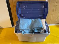 Open plastic storage bin with blue lid resting on floor, containing several folded blankets in light blue, beige, and plaid patterns.