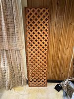 Full view of the cedar trellis panel leaning against a wood paneled wall with curtain on left and light cord near base, showing overall size and condition.