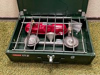 Front view of open Coleman 425 stove showing two burners, red fuel tank, metal funnel, and cooking grate inside dark green metal case.