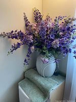 Wide shot of the large faux flower arrangement in a white ceramic vase placed on folded rugs or towels against a corner wall under natural light from a nearby window.