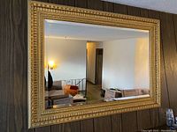 Full view of the gilt-frame wall mirror hanging on a wall with dark wood paneling. Reflects part of a living room area with furniture and boxes.