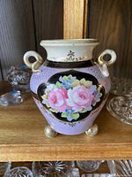 Front view of hand painted Nippon vase showing floral design with pink roses and blue flowers on a black oval background, light purple body, gold detailing, and two loop handles.