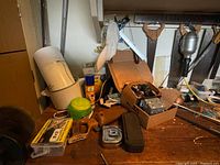 Wide view of a workbench showing various vintage handsaws hanging above and a box of hardware parts with loose tools and tape measure on the bench.