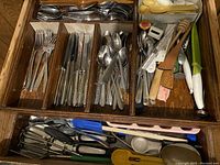 Drawer with assorted vintage flatware separated in compartments, including forks, knives, spoons, and a variety of kitchen utensils like wooden spoons, spatulas, and other cooking tools.