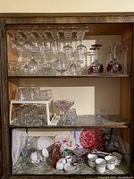 Full display of wooden cabinet shelving showcasing clear glass stemware on top shelf, smaller glass items and bowls on middle shelf, and white ceramic cups and saucers with decorative plates on bottom shelf.