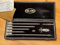 Top-down view of M&W Moore & Wright micrometer set inside black case showing micrometer head, 5 extension rods, one locking nut and adjustment tool, with M&W logo in gold on case lining.