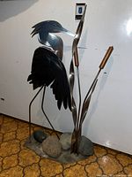 Front and side views of the metal heron garden sculpture showing painted metal bird, cattail leaves with wooden tops, and rock base.