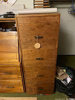 Full view of the tall wooden filing cabinet showing four compartments with metal handles, drop-front compartment tied with string and wooden disc on top.