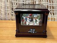 Front view of the wooden music box with a glass window showing the winter scene figurines inside and a decorative bow on the base.