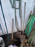 Three long-handled yard tools leaning against a textured wall: 2 shovels with metal spade blades and wooden handles, and one pickaxe with dual metal heads and wooden handle. Garden hoses seen coiled nearby.