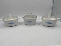 Lot of three white Corningware deep dishes with blue cornflower pattern, each with glass lid.