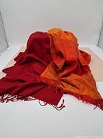Two scarves displayed together, one solid red and one orange with pattern, both with fringed edges.