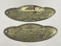 Photo showing both brass restroom plaques 'GENTLEMEN' and 'LADIES' side by side from top view to display wear and condition.