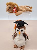 Photo of the two Ty Beanie Babies: a lion plush on top and a graduation penguin plush below on a clear stand.
