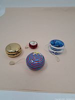 Four vintage yo-yos on flat surface showing various colors and materials including brass, red, blue transparent plastic and multicolor swirl plastic.