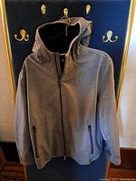 Front view of gray Tommy Hilfiger men's hooded coat hanging, showing zippered pockets and Tommy Hilfiger logo on chest.