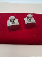 Two vintage metal bottles on red cloth, showing front and top views, highlighting shape, metal finish and capped tops.