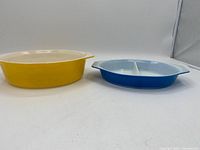 Yellow oval Pyrex baking dish and blue round divided Pyrex dish photographed side by side on a white surface.