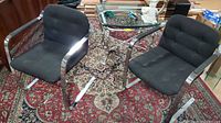 Both black upholstered mid-century modern cantilever chairs shown together on patterned carpet. Chrome metal cantilever frames and padded fabric cushions visible.